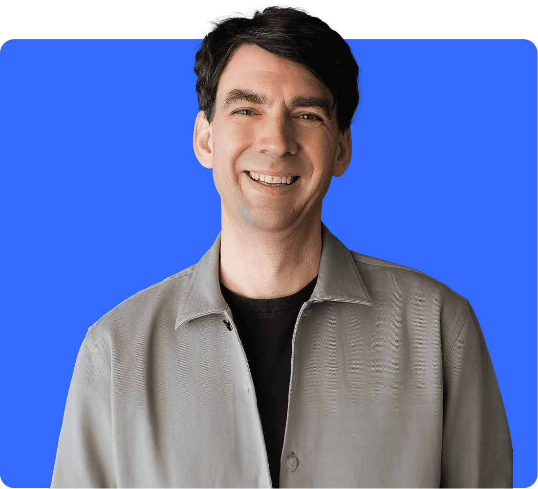 Portrait de Matthias « Matze » Henze, directeur général et fondateur de Jimdo, sur un fond bleu. Il porte une chemise grise et sourit à l'objectif.